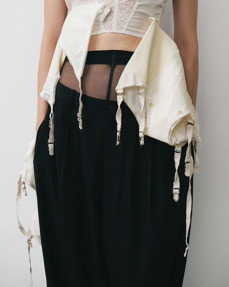 Fashion shot of a woman wearing an ivory lace corset with garter straps and long black trousers against a plain, light background.
