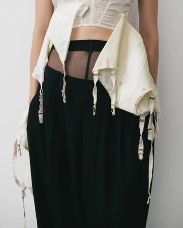 Fashion shot of a woman wearing an ivory lace corset with garter straps and long black trousers against a plain, light background.