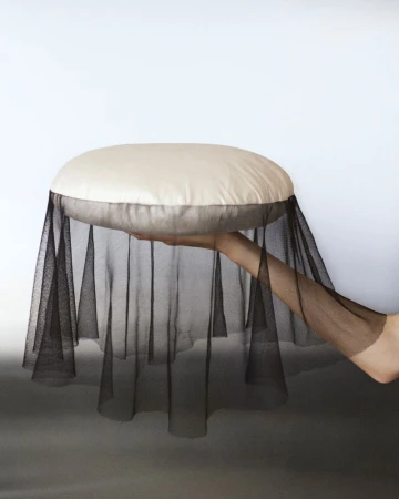 A person holds a round, cushioned object covered in black tulle, set against a plain white and gray background in a studio setting.