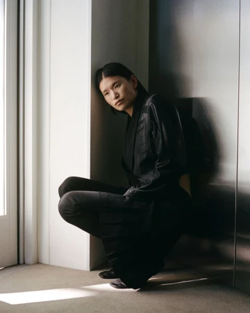 Young woman in a black outfit squats indoors in a corner with natural light, facing forward with a neutral expression in a portrait.