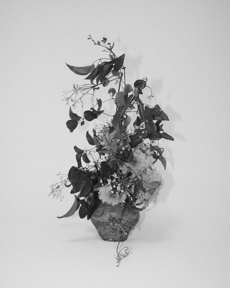 Black and white image of a floral arrangement in a vase. Various types of flowers and leafy vines fill the frame.