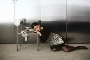 Model Cecilia Wu reclines on the floor, resting her head on a minimalist metal chair against a brushed stainless steel wall.