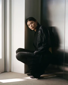 Model Cecilia Wu crouching in an all-black tailored ensemble against a white architectural pillar and metallic wall panel.