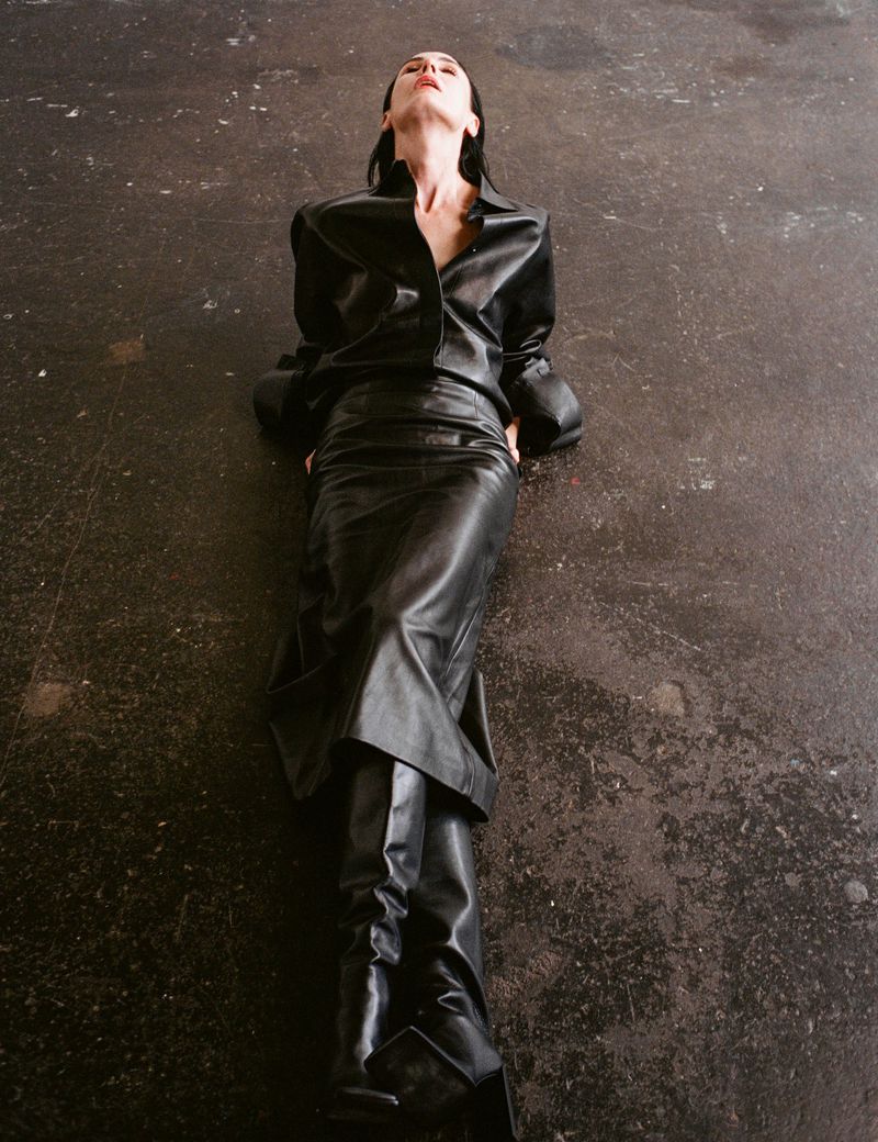 Woman in black leather shirt, skirt, and boots lying on grey concrete floor. Fashion editorial portrait with dramatic lighting and minimalist aesthetic.