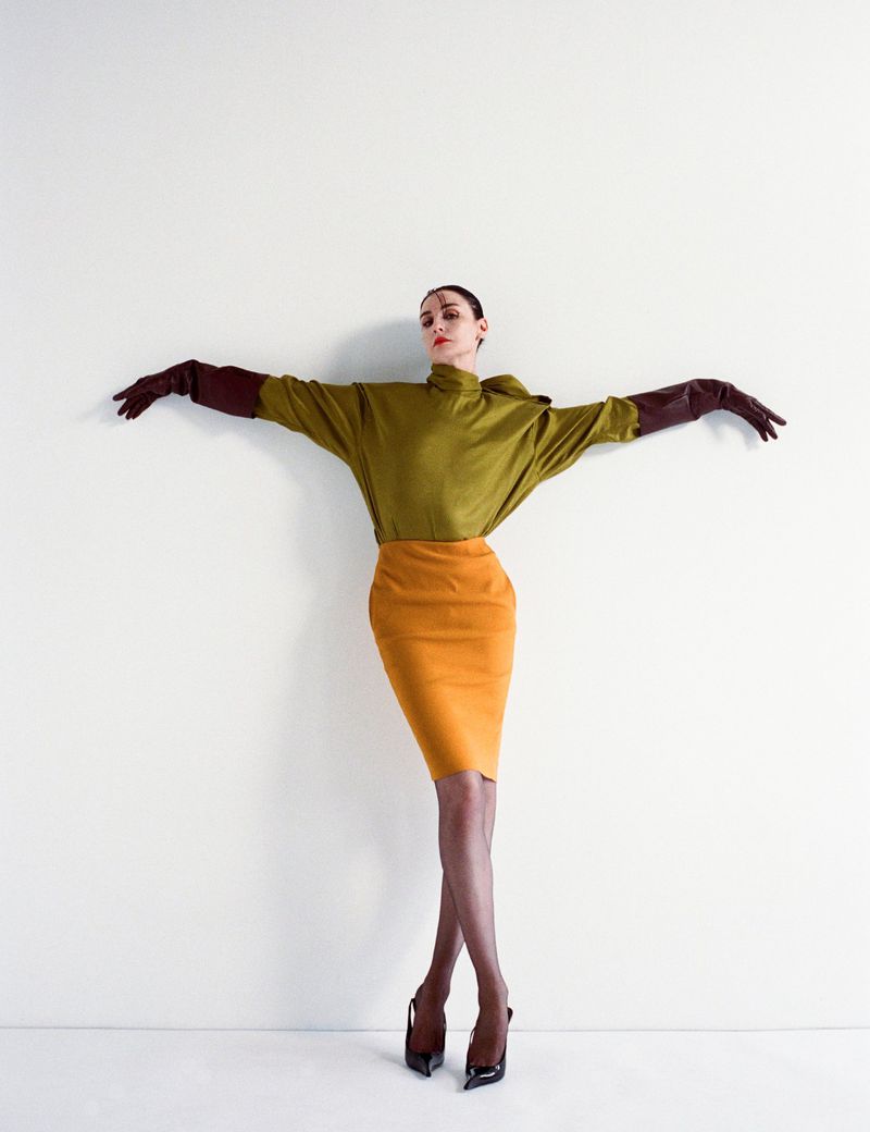 Adult female fashion model in olive top, orange skirt, gloves and heels poses against a white wall in a studio setting.