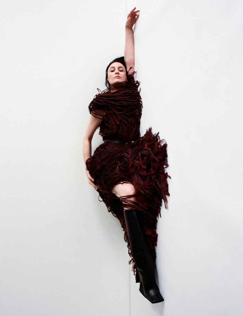 Fashion shot of a woman in a feathered dress and boots, lying down against a white wall, with one arm raised, captured in a portrait.