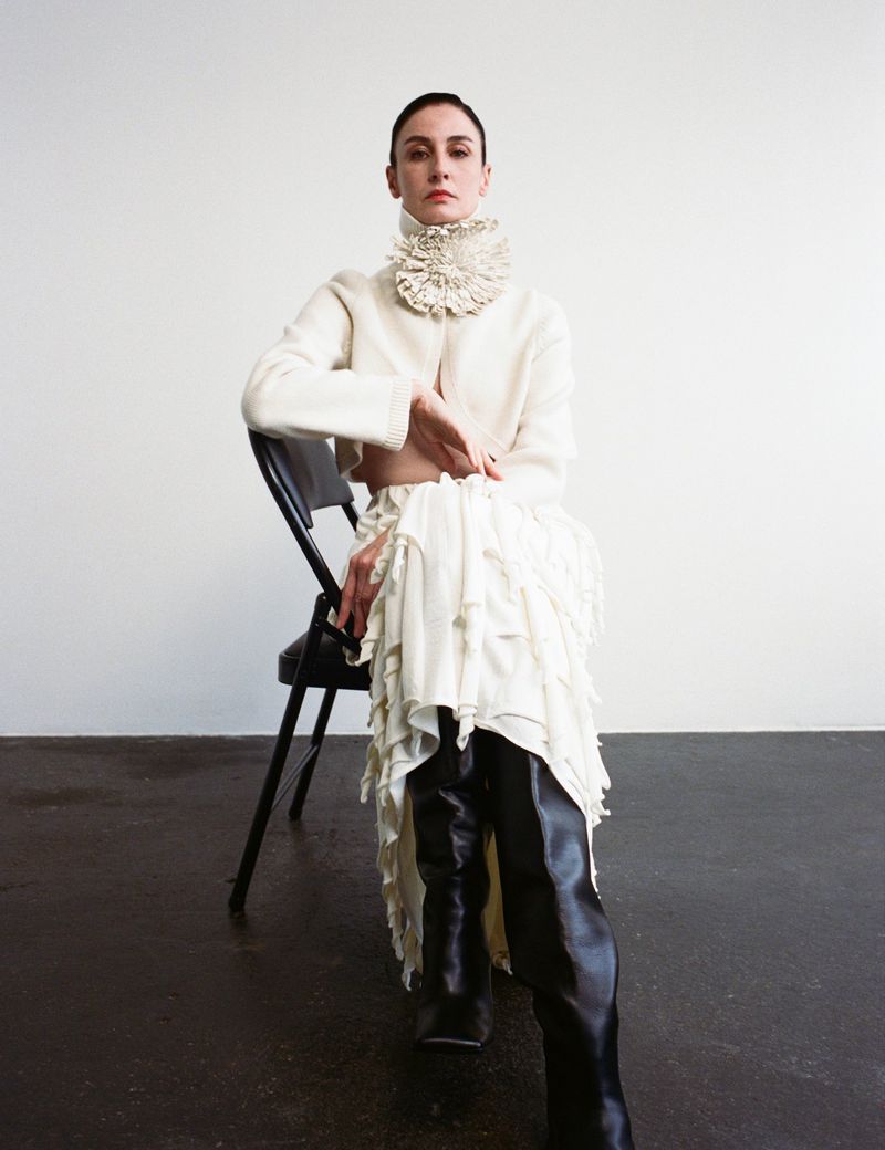 Elegant woman seated wearing ruffled collar and skirt with black thigh-high boots against white and dark gray background.