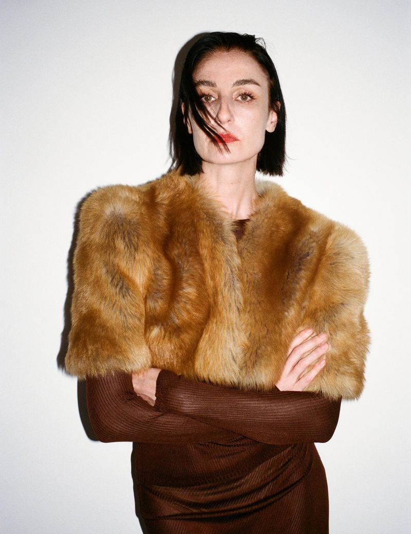 Portrait of a woman wearing a brown faux fur stole and a brown dress, facing the camera with arms crossed in a photo studio.