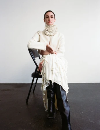 Erin O'Connor seated on a black chair wearing a cream textured collar sweater and black boots against a neutral background.