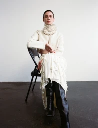 Erin O'Connor seated on a black chair wearing a cream textured collar sweater and black boots against a neutral background.