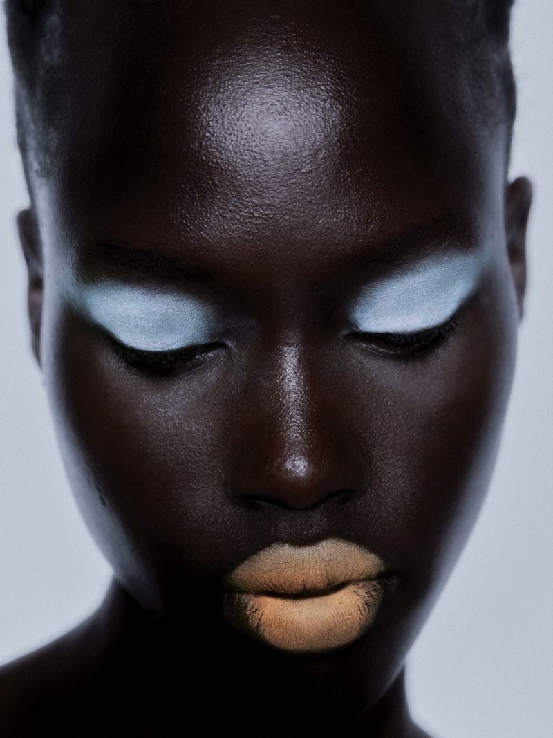 Close-up beauty shot of a dark-skinned adult female with soft, light blue eyeshadow and light peach-colored lipstick against a neutral backdrop.