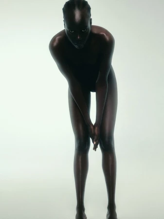 Full-body shot of a slender, dark-skinned woman in a studio, bending forward with hands together, exhibiting a contemplative and intimate pose.