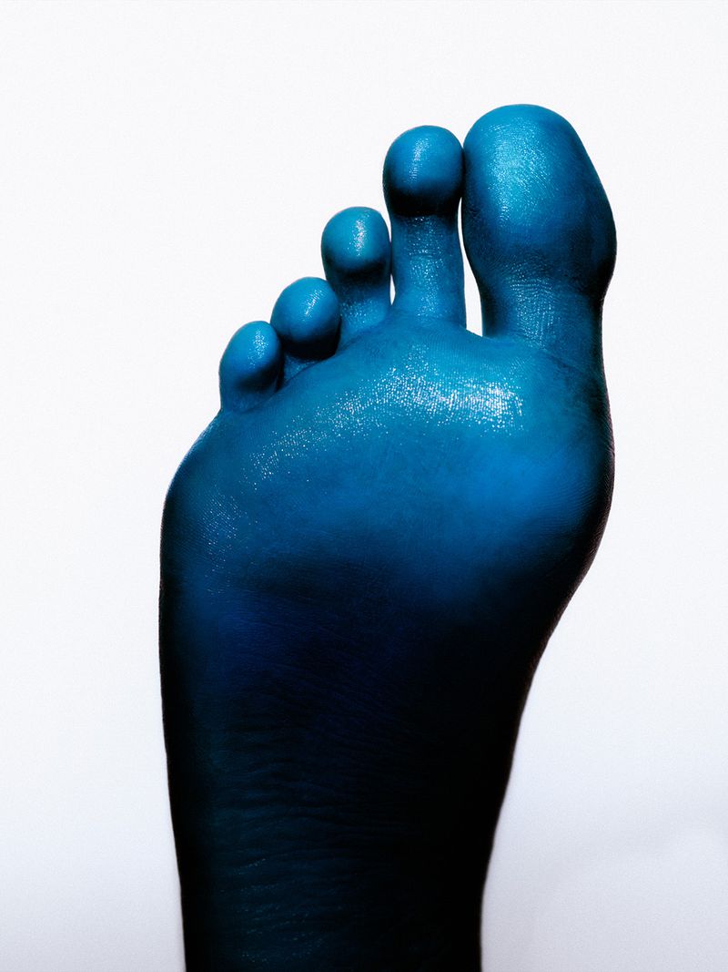 Macro photo of an adult foot painted glossy blue against a bright white background showcasing body art, skin texture, and toe details.