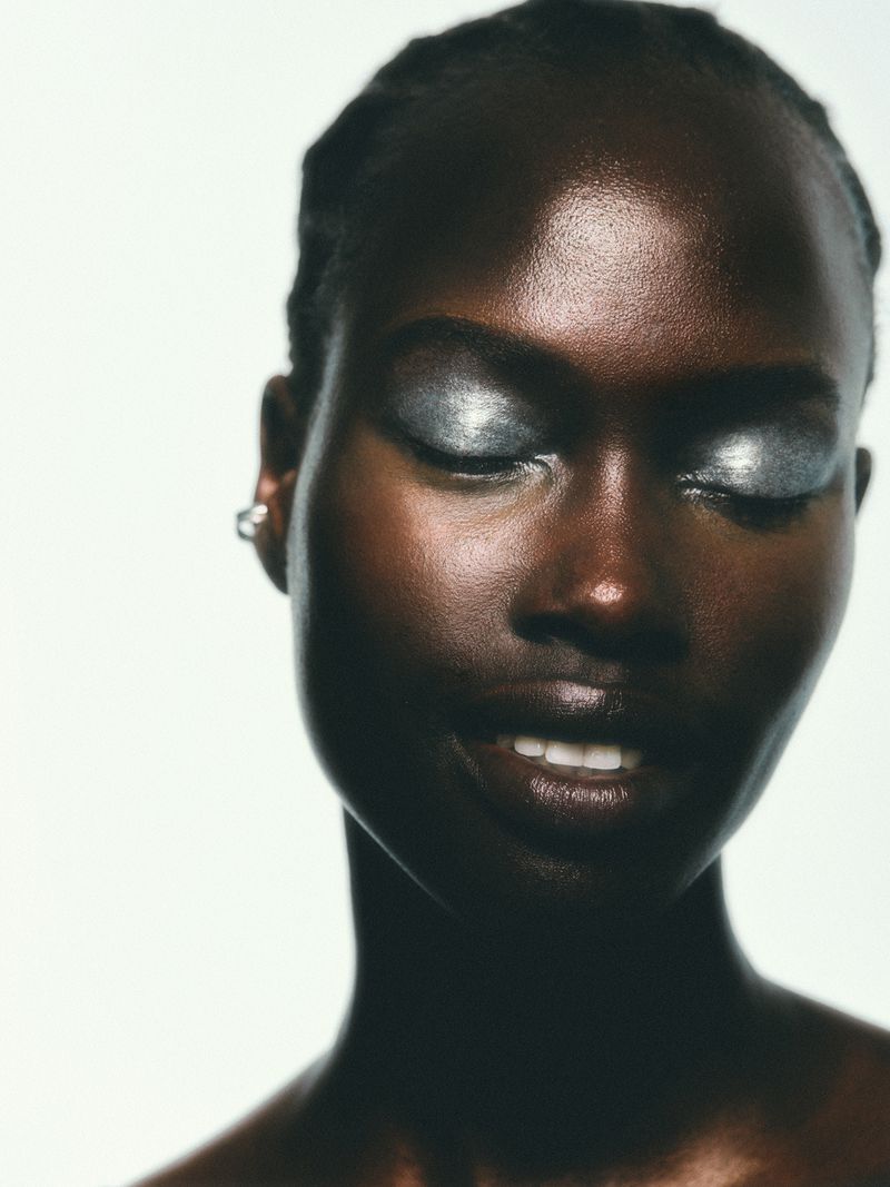 Headshot of a dark-skinned woman with silver eye makeup, lips slightly parted, shot against a white background in a beauty context.