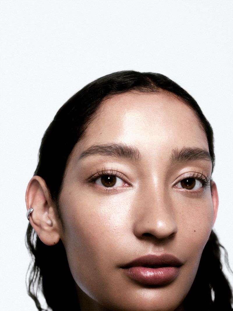 Close-up portrait of a woman with dark hair pulled back, showcasing smooth skin, natural makeup, and a silver earring against a bright white background.