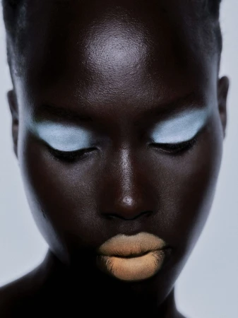 Close-up beauty portrait of a Black woman with pale powder blue eyeshadow and nude matte lipstick against a neutral backdrop.