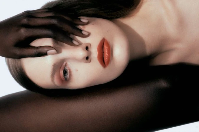 Close-up beauty portrait of a woman with orange lipstick and a dark hand resting across her forehead against a white background.
