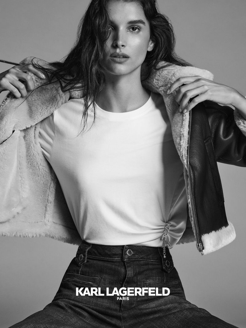 Black-and-white fashion portrait of a woman in a white T-shirt, black shearling jacket, and wide-leg jeans, posing against a grey background.