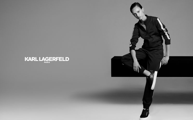 Monochrome fashion shot of an adult female model in a tracksuit sitting on a black block in a studio with a gray background.