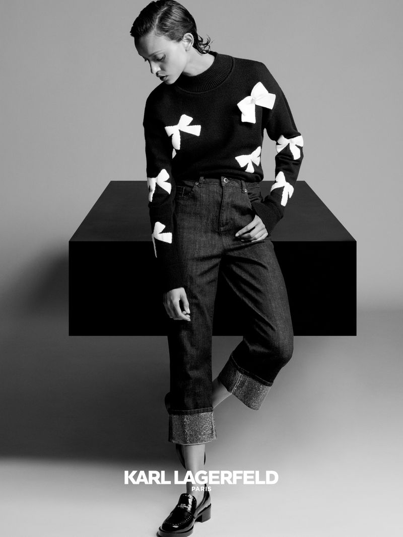 Monochrome studio shot of a female model in a dark sweater with white bows and dark cuffed denim jeans posing in front of a black table.