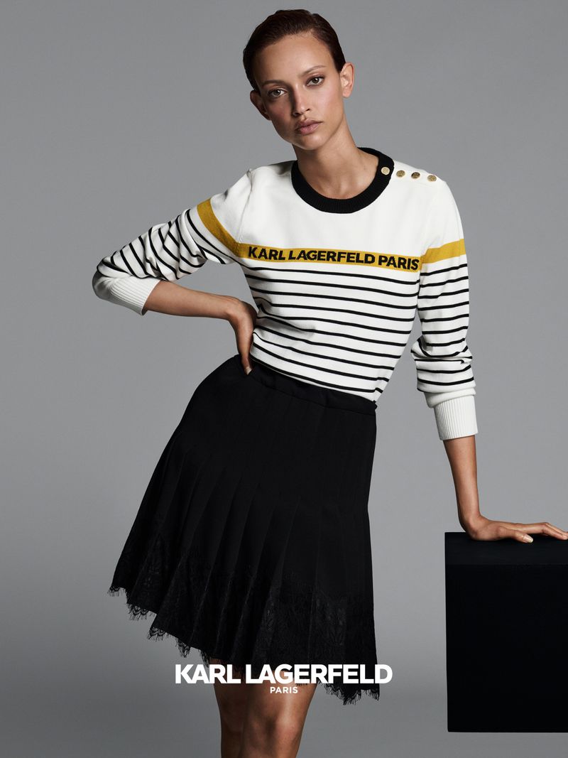 Elegant woman poses indoors wearing a Karl Lagerfeld striped sweater and a black lace skirt against a gray backdrop in a fashion shot.