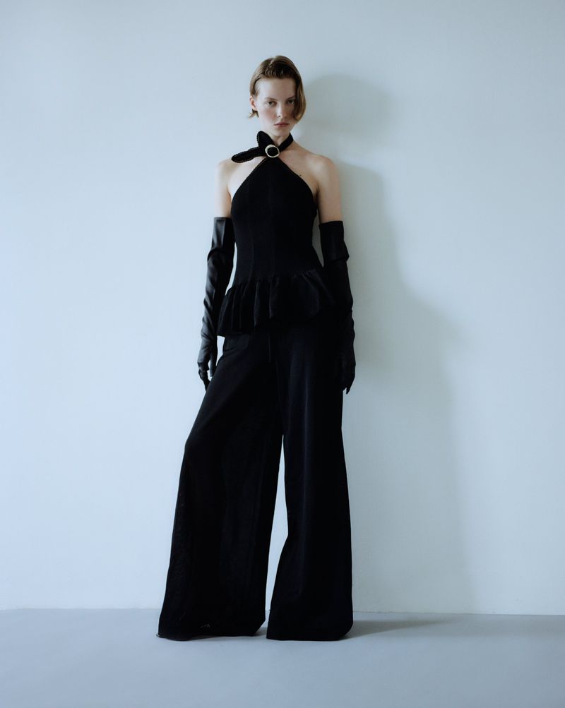 Fashion model in black halter top and wide-leg pants poses against a pale blue background in a studio setting, accessorized with long gloves.