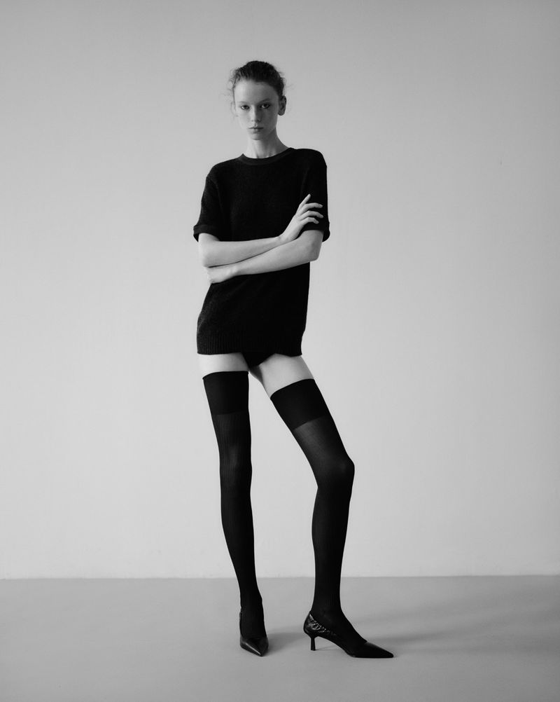 Studio shot of an adult woman in dark top and stockings, arms crossed, looking at camera against a neutral gray background.