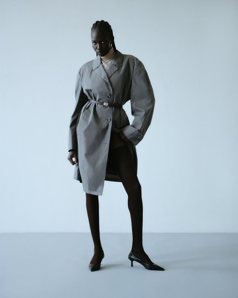 Black female model wearing a grey belted trench coat, black tights, and pointed heels against a studio background.