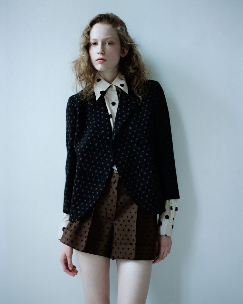Model standing against a plain pale background wearing a dark patterned blazer and brown patterned shorts.