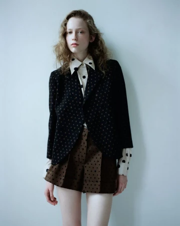 Model standing against a plain pale background wearing a dark patterned blazer and brown patterned shorts.