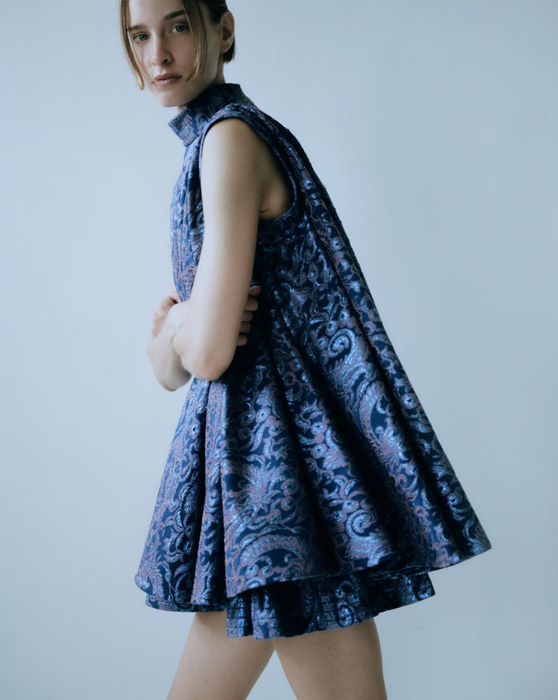A stylish young woman poses in an elegant patterned blue dress, arms crossed, against a clean, light backdrop.