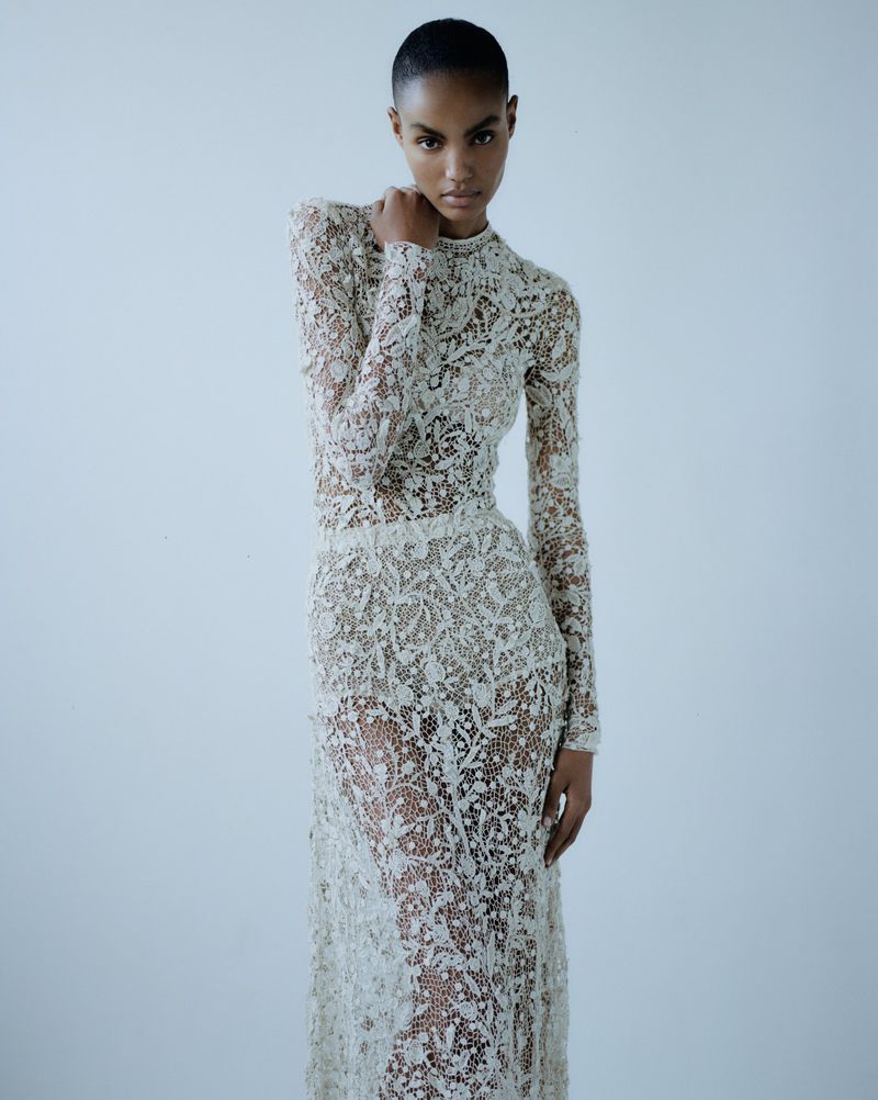 Elegant Black model poses in a full-length, long-sleeved, off-white, floral lace gown against a clean, light backdrop.