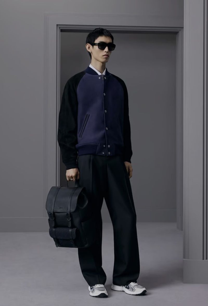 Man in a blue and black jacket, sunglasses, and black pants, holding a backpack, in front of gray wall and doorway.