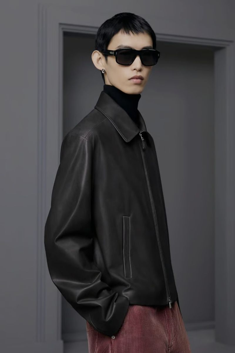 Stylish man in leather jacket and sunglasses posing against grey background. Corduroy pants, black turtleneck, and earring.