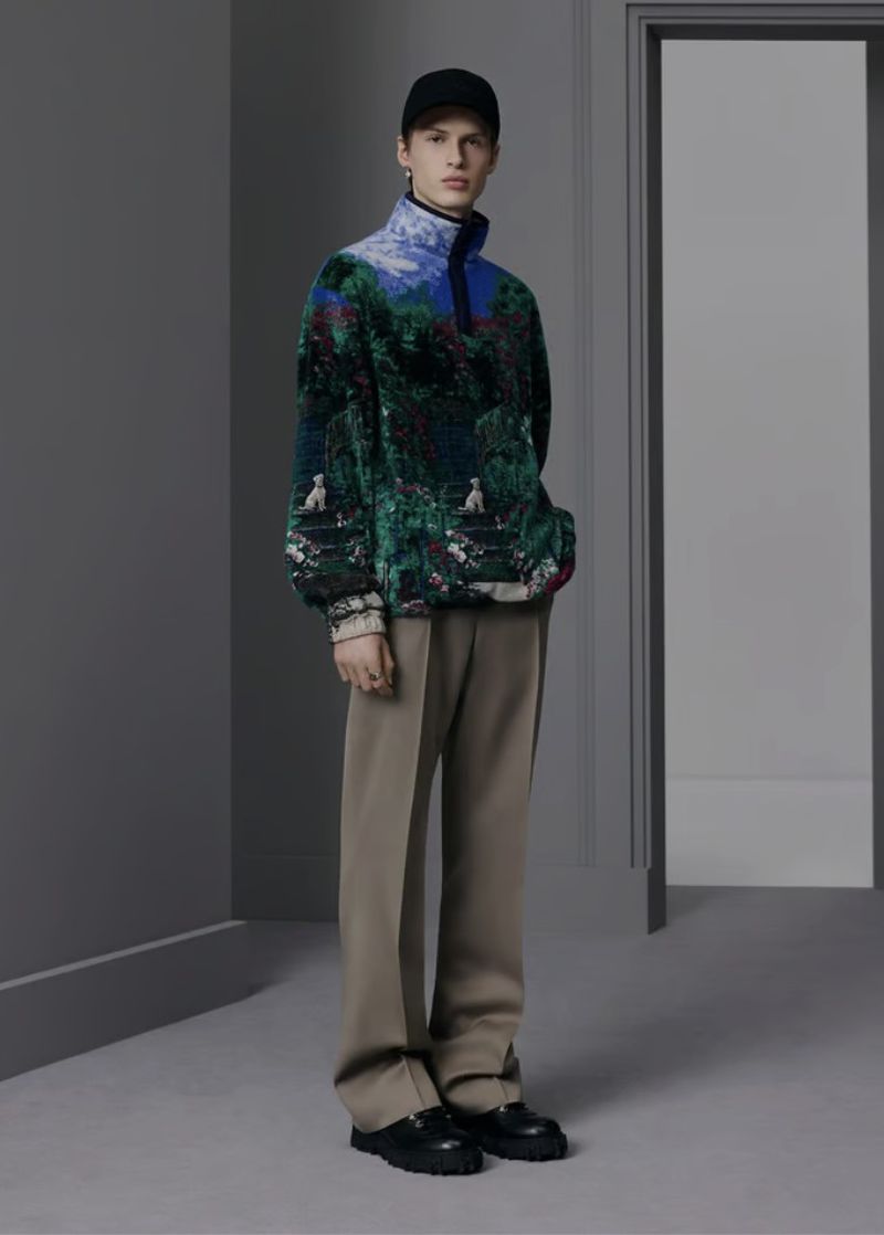 Full-length studio shot of an adult male model wearing a colorful graphic sweater, taupe pants, black cap and platform loafers.