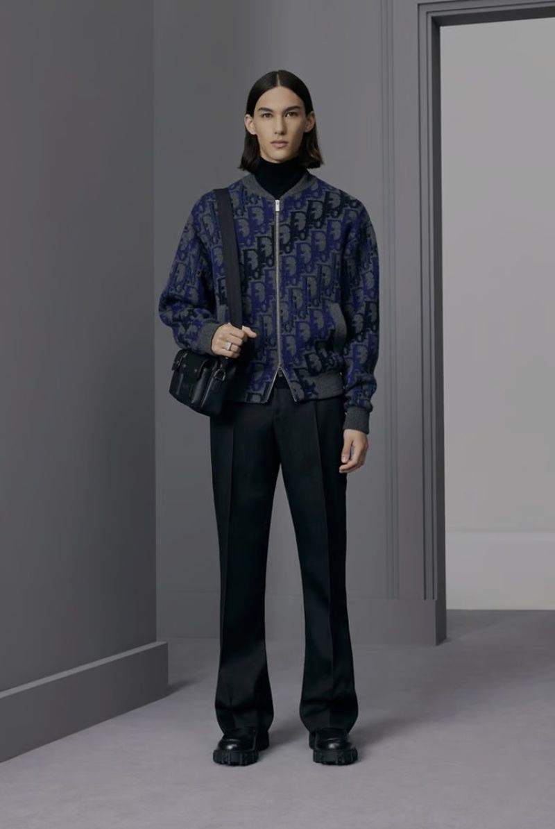 Full shot of a young adult male in a printed bomber jacket and black pants, standing indoors with a shoulder bag, looking at the camera.
