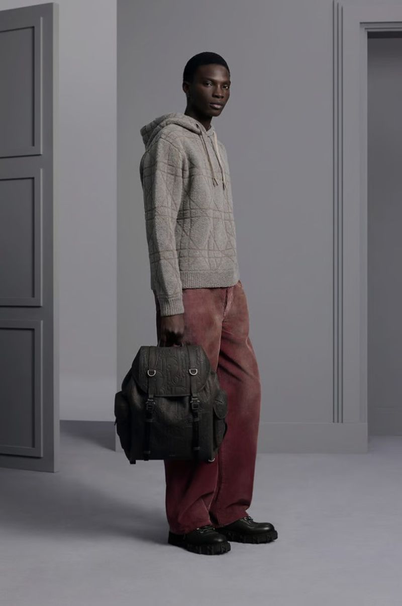 A dark-skinned man models a grey hoodie and a black backpack in a studio setting, wearing wide-leg pants and black shoes.