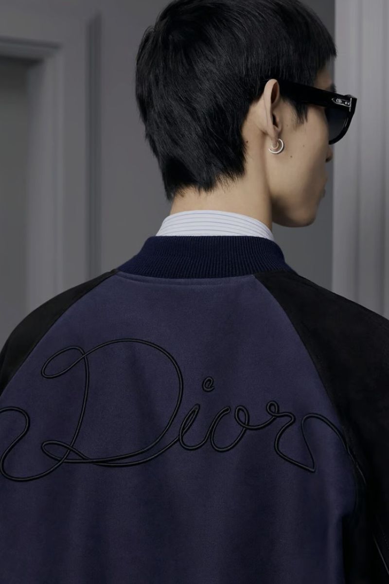 Male fashion model wears a bomber jacket with the 'Dior' logo on the back, paired with sunglasses and an earring, in a studio setting.