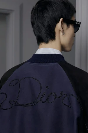 Male fashion model wears a bomber jacket with the 'Dior' logo on the back, paired with sunglasses and an earring, in a studio setting.