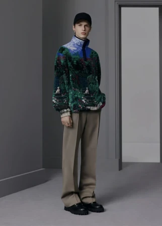 Model posing in a scenic landscape print fleece pullover, taupe wide-leg trousers, and a black cap in a grey architectural room.