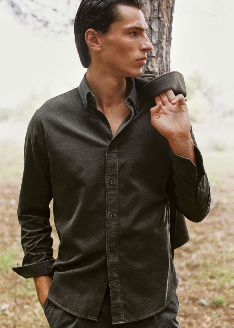 A man stands in a natural outdoor setting wearing a dark button-down shirt with a jacket slung over his shoulder in a portrait style.