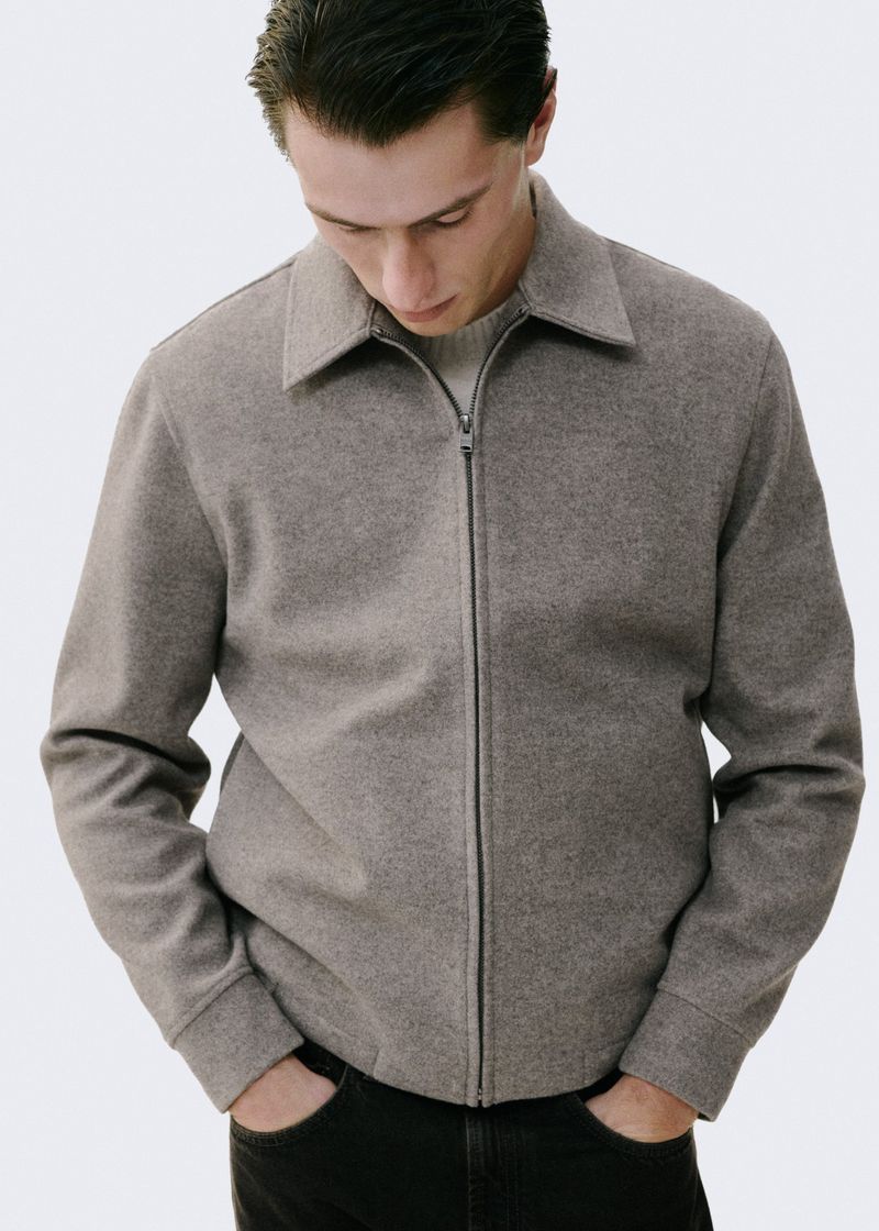 A light-skinned man in a casual taupe jacket with a zipper, set against a bright background, embodies minimalist fashion.