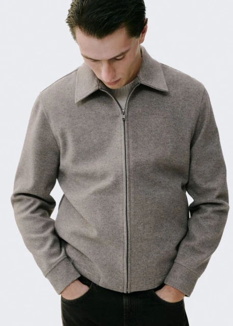 A light-skinned man in a casual taupe jacket with a zipper, set against a bright background, embodies minimalist fashion.