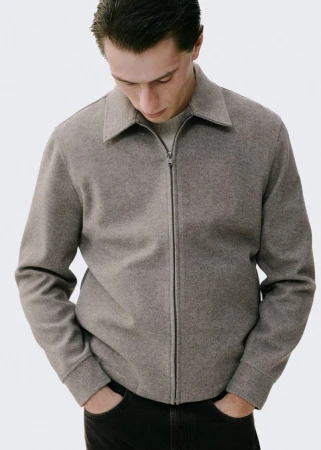 A light-skinned man in a casual taupe jacket with a zipper, set against a bright background, embodies minimalist fashion.