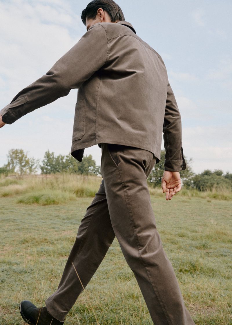 A man in a brown corduroy suit walks across a grassy field under a partly cloudy sky in a fashion or editorial style outdoor shot.