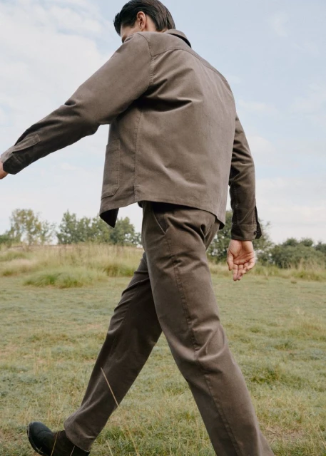 A man in a brown corduroy suit walks across a grassy field under a partly cloudy sky in a fashion or editorial style outdoor shot.