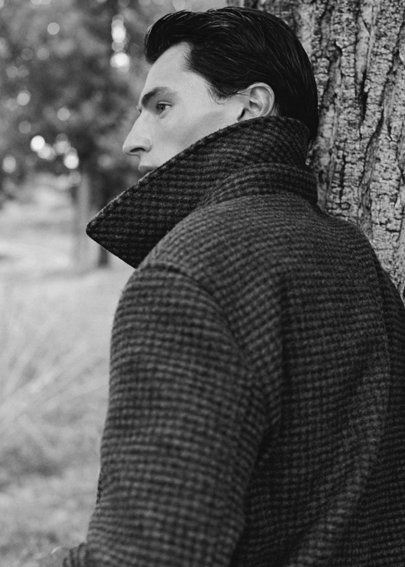 Black and white portrait of an adult male model in a patterned tweed coat leaning against a tree in an outdoor setting with bokeh.