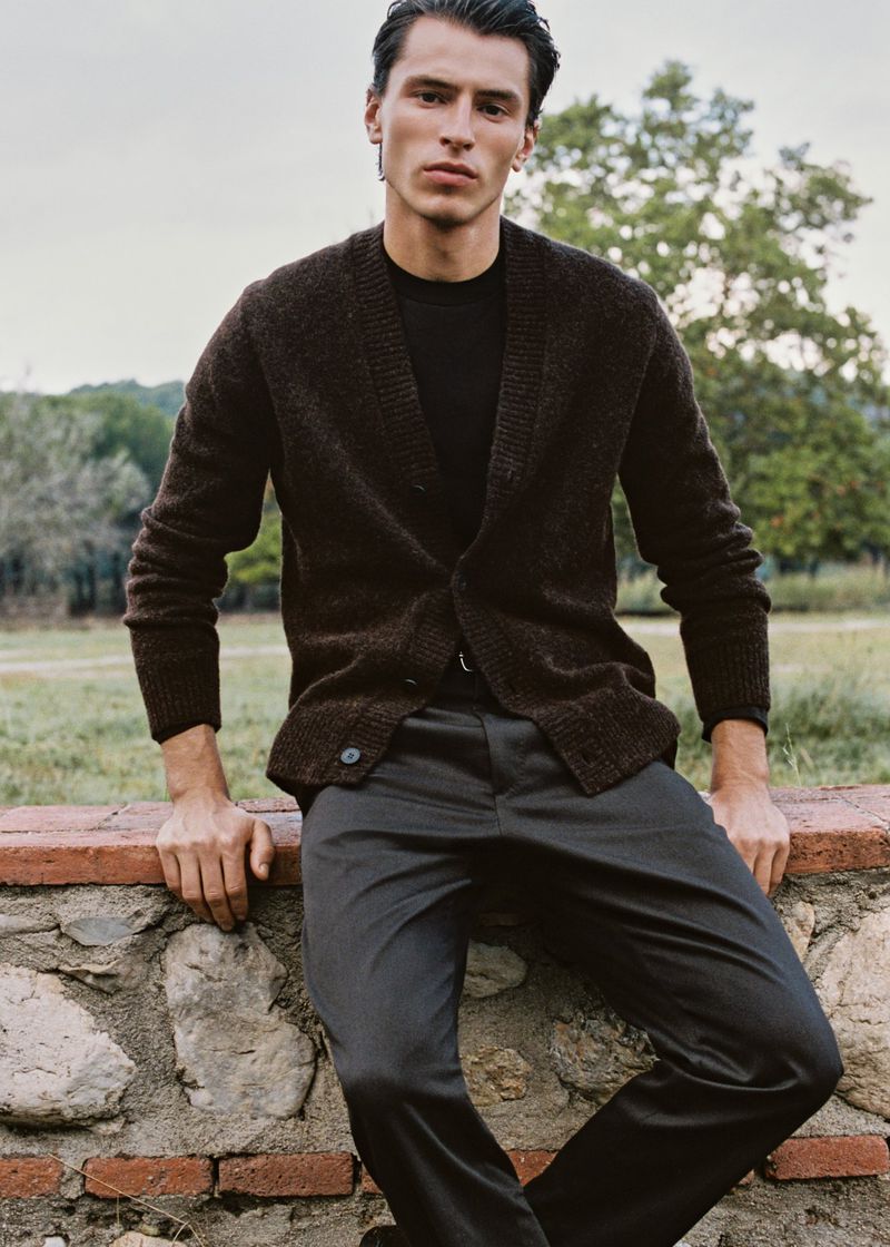 A man in a dark cardigan and pants leans against a low stone wall, posing outdoors with a backdrop of trees and sky in natural light.