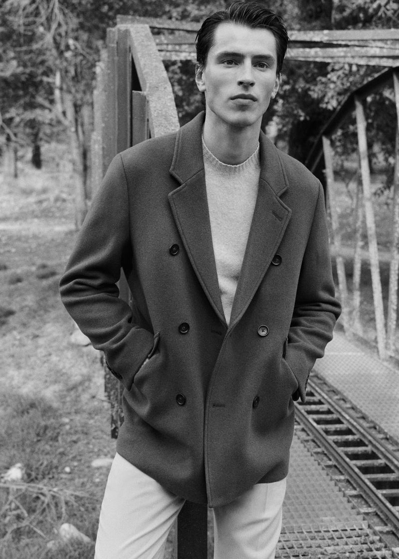 A grayscale portrait of a young adult male model in a coat and sweater, standing in front of a metal bridge and trees in an outdoor setting.