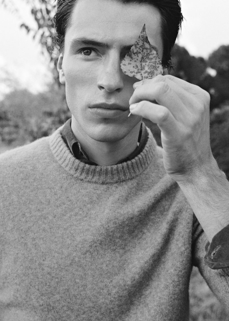 Black and white portrait of adult male in sweater holding a leaf over one eye in an outdoor setting with a blurred background.